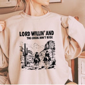 May include: A beige sweatshirt with a black graphic print of two cowboys riding horses in a desert landscape. The text "LORD WILLIN' AND THE CREEK DON'T RISE" is printed above the graphic.