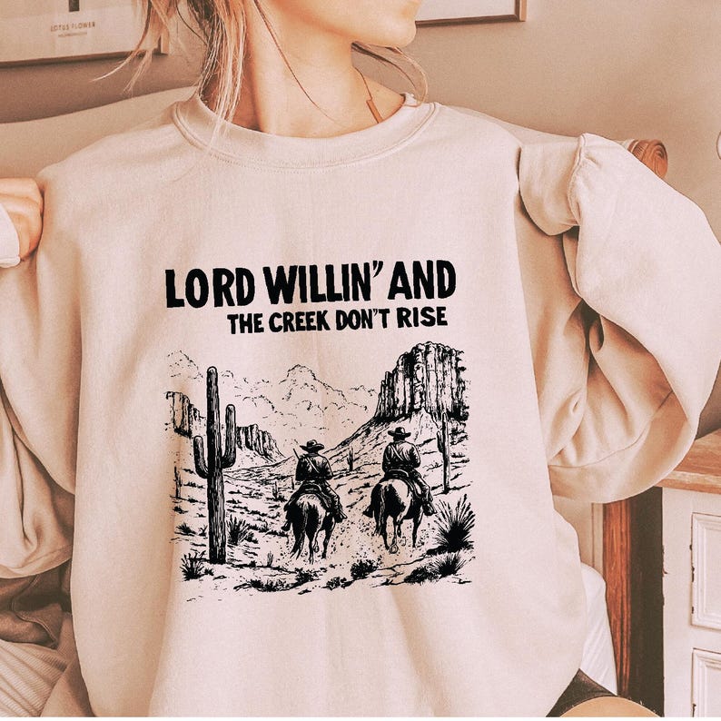 May include: A beige sweatshirt with a black graphic print of two cowboys riding horses in a desert landscape. The text "LORD WILLIN' AND THE CREEK DON'T RISE" is printed above the graphic.