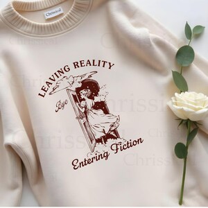 May include: A cream-colored sweatshirt with a brown graphic print of a woman stepping through a door into the clouds. The text reads "Leaving Reality Bye Entering Fiction".