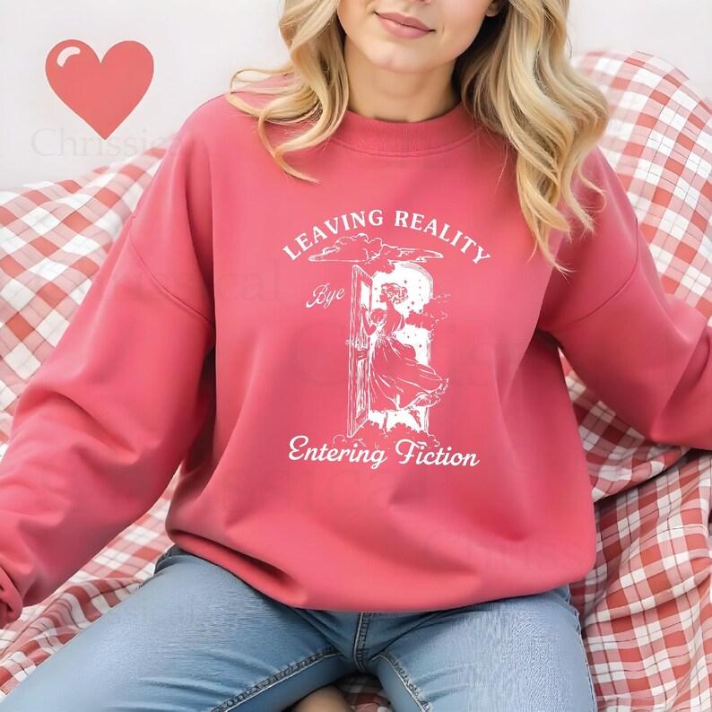 May include: A pink sweatshirt with a white graphic print that says "Leaving Reality Bye Entering Fiction". The graphic features a woman in a dress walking through a doorway.