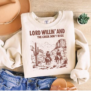 May include: A cream-colored sweatshirt with a brown graphic print of two cowboys riding horses in a desert landscape. The text "LORD WILLIN' AND THE CREEK DON'T RISE" is printed in brown on the sweatshirt.