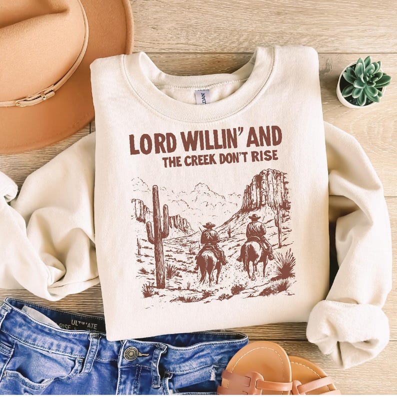 May include: A cream-colored sweatshirt with a brown graphic print of two cowboys riding horses in a desert landscape. The text "LORD WILLIN' AND THE CREEK DON'T RISE" is printed in brown on the sweatshirt.
