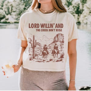 May include: A cream colored t-shirt with a brown graphic print of two cowboys riding horses through a desert landscape. The text "Lord Willin' and the Creek Don't Rise" is printed in brown on the shirt.