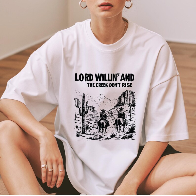May include: White t-shirt with a black graphic print of two cowboys riding horses in a desert landscape. The text "LORD WILLIN' AND THE CREEK DON'T RISE" is printed above the graphic.