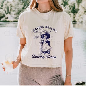 May include: A white t-shirt with a blue graphic print of a woman stepping through a door into a cloud. The text on the shirt reads "Leaving Reality Bye Entering Fiction".