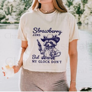 May include: A white t-shirt featuring a graphic of a raccoon wearing a cowboy hat and holding a gun and a glass. The text on the shirt reads "Strawberry Jams But My Glock Don't".
