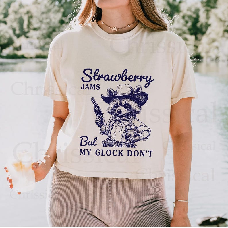 May include: A white t-shirt featuring a graphic of a raccoon wearing a cowboy hat and holding a gun and a glass. The text on the shirt reads "Strawberry Jams But My Glock Don't".