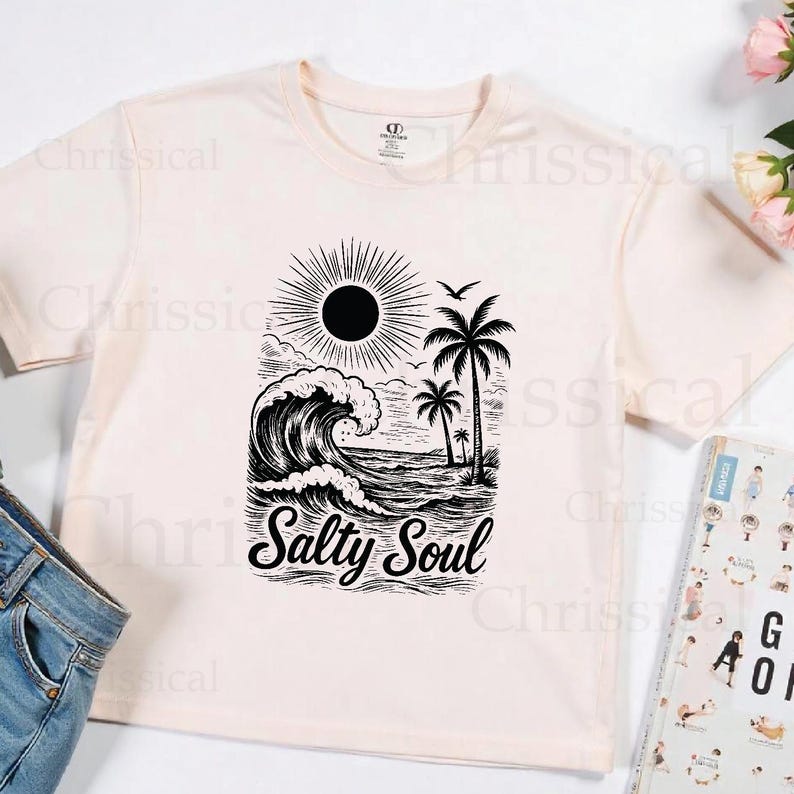 May include: A light pink t-shirt with a black and white graphic design of a wave, palm trees, and a sun. The text "Salty Soul" is printed in black below the graphic.