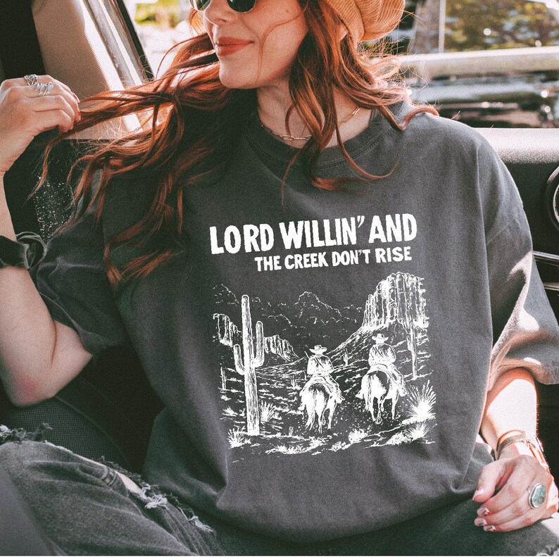 May include: A gray t-shirt with a white graphic print of two cowboys riding horses in a desert landscape. The text "Lord Willin' and the Creek Don't Rise" is printed above the graphic.