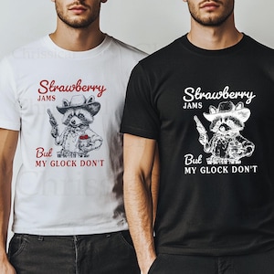 May include: Two t-shirts featuring a graphic of a raccoon wearing a cowboy hat and holding a gun. The text on the shirts reads "Strawberry Jams But My Glock Don't".