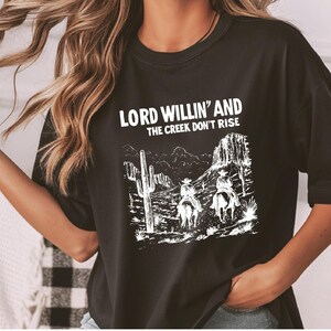 May include: Black t-shirt with a white graphic print of two cowboys riding horses in a desert landscape. The text "LORD WILLIN' AND THE CREEK DON'T RISE" is printed above the graphic.