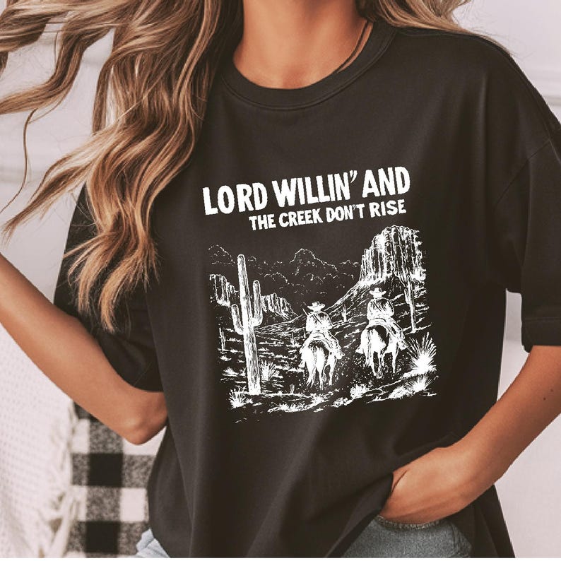 May include: Black t-shirt with a white graphic print of two cowboys riding horses in a desert landscape. The text "LORD WILLIN' AND THE CREEK DON'T RISE" is printed above the graphic.