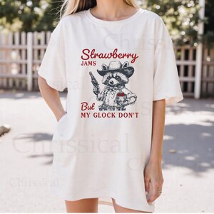 May include: White t-shirt featuring a graphic of a raccoon wearing a cowboy hat and holding a gun and a jar of strawberry jam. The text on the shirt reads "Strawberry Jams But My Glock Don't".