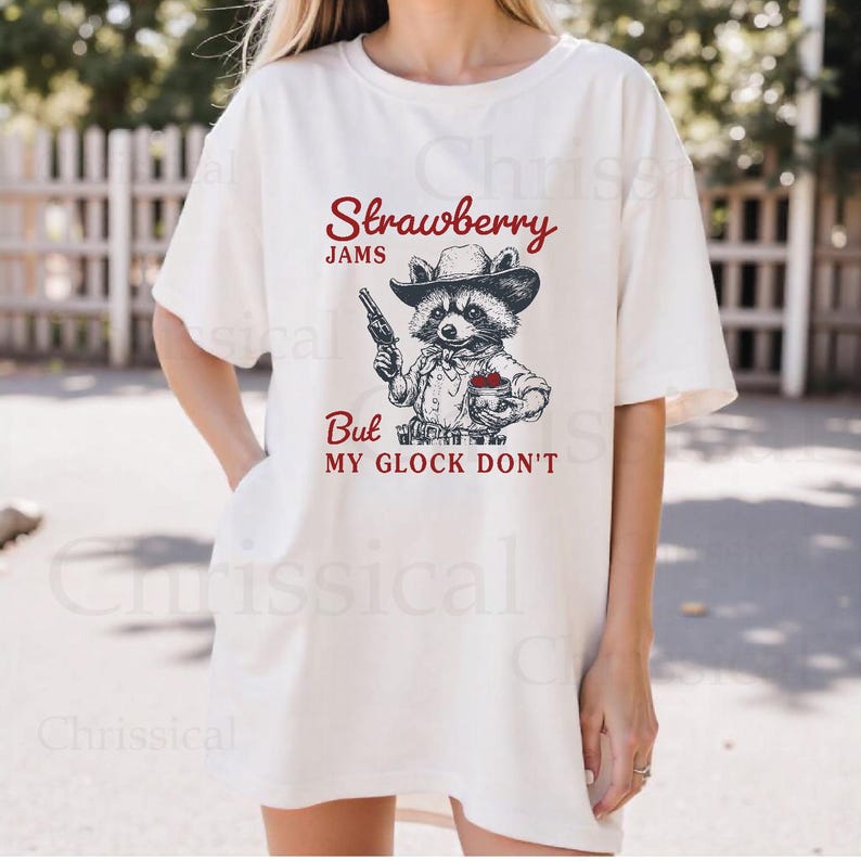 May include: White t-shirt featuring a graphic of a raccoon wearing a cowboy hat and holding a gun and a jar of strawberry jam. The text on the shirt reads "Strawberry Jams But My Glock Don't".