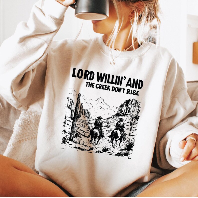 May include: A white sweatshirt with a black graphic print of two cowboys riding horses in a desert landscape. The text "LORD WILLIN' AND THE CREEK DON'T RISE" is printed on the sweatshirt.