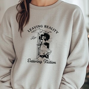 May include: A gray sweatshirt with a black graphic print of a woman stepping through a door with the text "Leaving Reality Bye Entering Fiction".