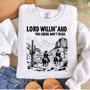 May include: A gray sweatshirt with a black graphic print of two cowboys riding horses. The text "Lord Willin' and the Creek Don't Rise" is printed on the sweatshirt.