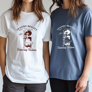 May include: Two white t-shirts with a graphic print of a girl stepping through a door. The text on the shirts reads "Leaving Reality Bye Entering Fiction".