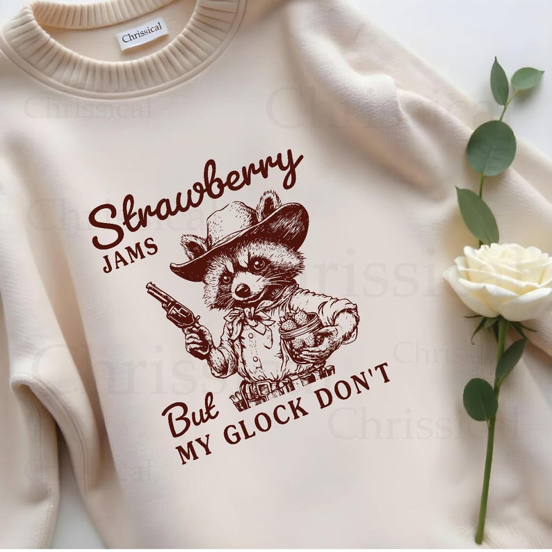 May include: A cream-coloured sweatshirt with a brown illustration of a raccoon wearing a cowboy hat and holding a gun and a basket of strawberries. The text "Strawberry Jams But My Glock Don't" is printed on the sweatshirt.