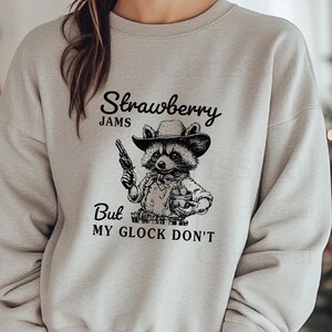 May include: A grey sweatshirt featuring a black and white illustration of a raccoon wearing a cowboy hat and holding a gun and a basket of strawberries. The text "Strawberry Jams But My Glock Don't" is printed on the sweatshirt.