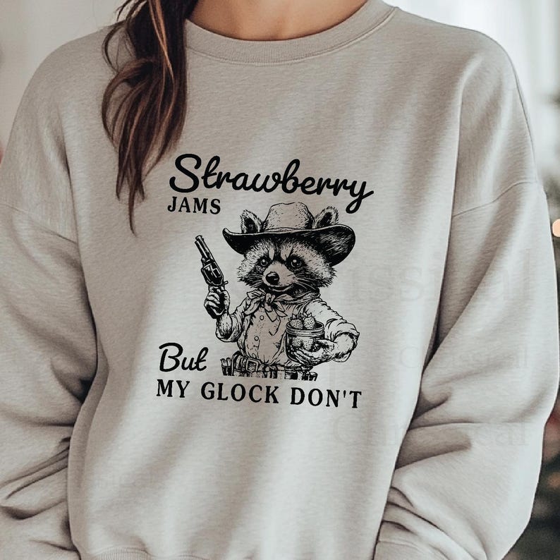 May include: A grey sweatshirt featuring a black and white illustration of a raccoon wearing a cowboy hat and holding a gun and a basket of strawberries. The text "Strawberry Jams But My Glock Don't" is printed on the sweatshirt.
