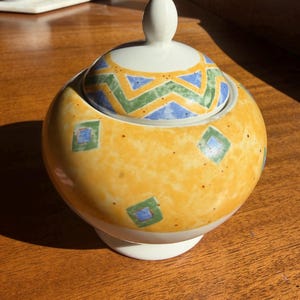 May include: A ceramic sugar bowl with a yellow exterior, decorated with blue and green geometric patterns. The lid features a white knob and a matching design. The bowl sits on a white base, perfect for serving sugar or other condiments.