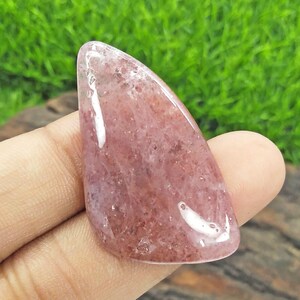 May include: A polished, teardrop-shaped gemstone with a translucent, reddish-pink hue. The stone has a smooth, glossy surface with subtle internal patterns and inclusions. The gemstone is held in a hand against a backdrop of green grass.