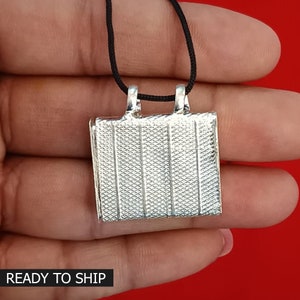 May include: Silver pendant with a square shape and a textured surface. The pendant is attached to a black cord.