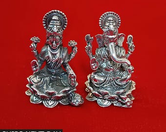 Op de afbeelding: Twee zilverkleurige beelden van de hindoeïstische goden Lakshmi en Ganesha op een rode achtergrond. Lakshmi, links, houdt lotusbloemen vast. Ganesha, rechts, heeft een olifantenkop. De beelden zijn gedetailleerd en sierlijk.