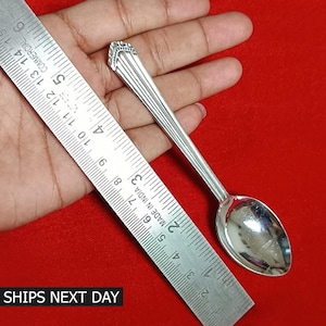 May include: A silver teaspoon with an ornate handle. The spoon is approximately 5 inches long and is measured against a ruler.