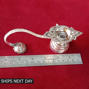 Puede incluir: Lámpara de aceite plateada con un diseño ornamentado. La lámpara tiene un asa curva y una pequeña base redonda. La lámpara mide aproximadamente 15 cm de largo.