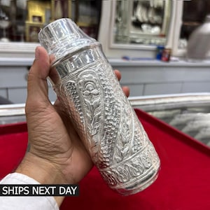 May include: A cylindrical silver cocktail shaker featuring an intricate embossed floral pattern and a textured hammered finish. The metal vessel has a tapered top section and a smooth, polished rim, showcasing detailed craftsmanship throughout the body.