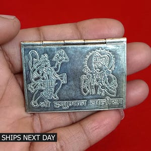 May include: A silver metal box with a hinged lid. The box features an engraved image of two Hindu deities, Hanuman and Ram, with the text "श्री हनुमान चालीसा" in Hindi.