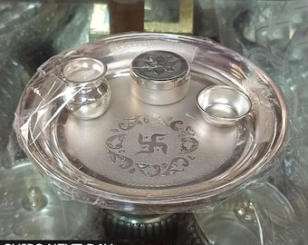 Può includere: Un thali d'argento, o piatto cerimoniale, con un disegno di svastica al centro. Il thali è circondato da tre ciotole d'argento più piccole, una con un coperchio.