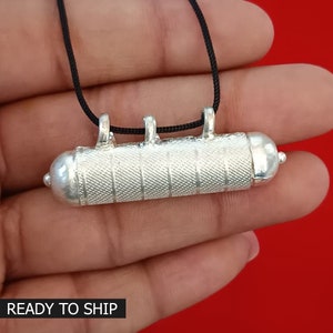May include: Silver pendant necklace with a cylindrical charm. The charm has a textured surface and is attached to a black cord.
