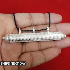 May include: A silver cylindrical pendant with a textured surface and three loops for attaching a cord. The pendant is suspended from a black cord.