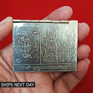 May include: A silver-colored, hinged, rectangular box with three engraved images of Hindu deities. The box has the text "श्री हनुमान ज्वालीसा" engraved on the bottom.