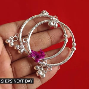 May include: Two silver baby bangles with a decorative pattern and small bells hanging from the bottom. The bangles are on a hand.