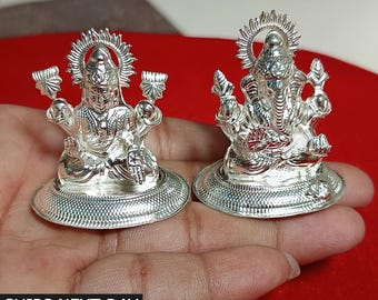 Op de afbeelding: Twee zilveren beelden van hindoeïstische goden, Lakshmi en Ganesha, op een hand. De beelden staan op ronde bases met ingewikkelde ontwerpen. De beelden zijn beide naar voren gericht en hebben een halo rond hun hoofd.