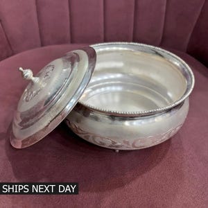 以下が含まれることがあります： 銀色の丸いサービングボウルと蓋。蓋は少し開いており、ボウルの内部が見えます。ボウルには装飾的なビーズの縁と花の彫刻があります。「SHIPS NEXT DAY」というテキストが下部に表示されています。