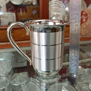 May include: A silver-colored metal mug with a handle. The mug has a brushed finish and a band of horizontal lines around the middle.