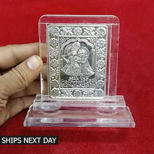 May include: A silver-colored religious figurine of Krishna and Radha, with intricate details, is displayed in a clear acrylic stand. The figurine is set against a red background.
