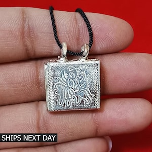 May include: Silver pendant necklace with a square charm featuring an intricate design of a deity riding a mythical creature. The pendant is attached to a black cord.