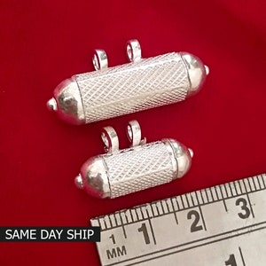 May include: Two silver, cylindrical, decorative pendants with a textured surface. The pendants are approximately 1 inch and 2 inches long. A ruler is in the background for size reference.