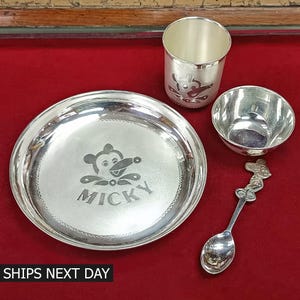 May include: A silver baby feeding set with a Mickey Mouse design. The set includes a plate, bowl, cup, and spoon.