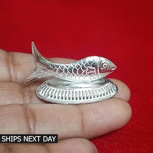 May include: A silver fish figurine with detailed scales and fins, resting on a decorative, round base. The figurine is held in a hand against a red background. The text "SHIPS NEXT DAY" is visible at the bottom.