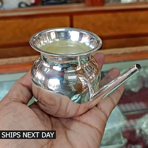 May include: A silver neti pot with a spout. The pot is round and has a smooth, shiny surface. It is filled with a clear liquid.