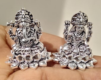 Op de afbeelding: Twee zilverkleurige beeldjes van de hindoe-goden Ganesha en Lakshmi, zittend op lotusbloem-bases. De beeldjes zijn gedetailleerd en ingewikkeld, met de goden afgebeeld in traditionele poses.