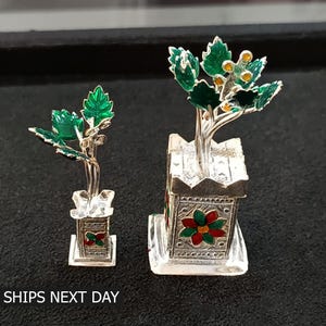 May include: Two silver miniature potted plants with green leaves and red and green flowers on the pots. The larger plant is on the right and the smaller plant is on the left.