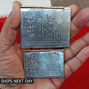 May include: Two silver folding boxes with engraved images of Hindu deities. The top box has three images, the bottom box has two images. The text on the boxes reads "Shri Hanuman Chalisa".